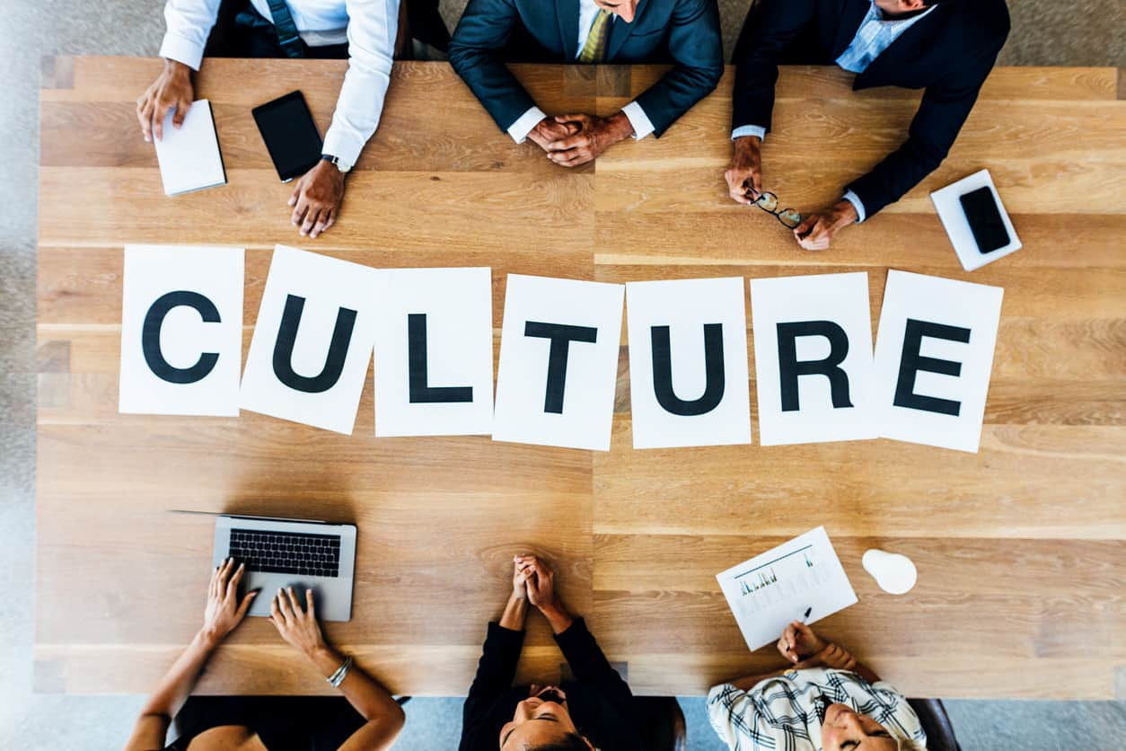 Two sets of three people seated on opposite sides along a table’s length, with the word culture written in different sheets of paper in the table space between them.