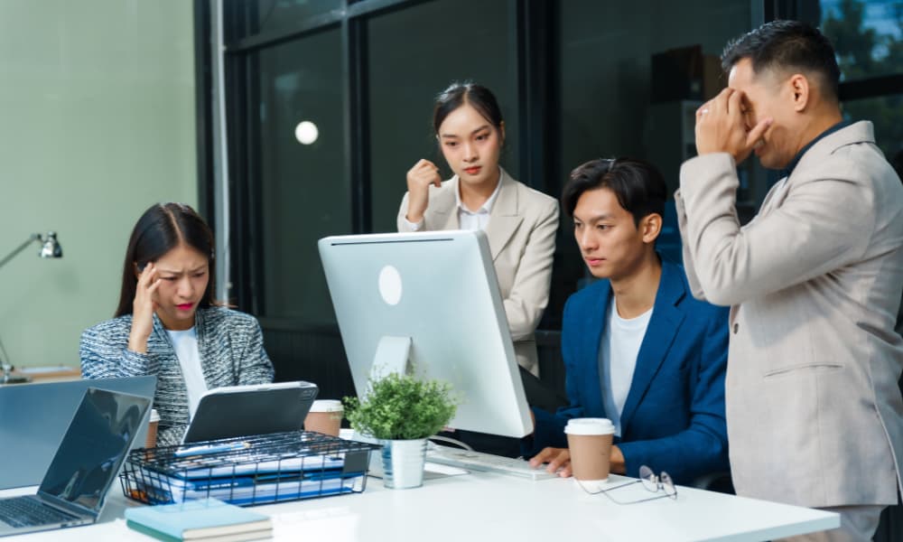 In an office, stressed Asian colleagues discuss financial failures at a desk.