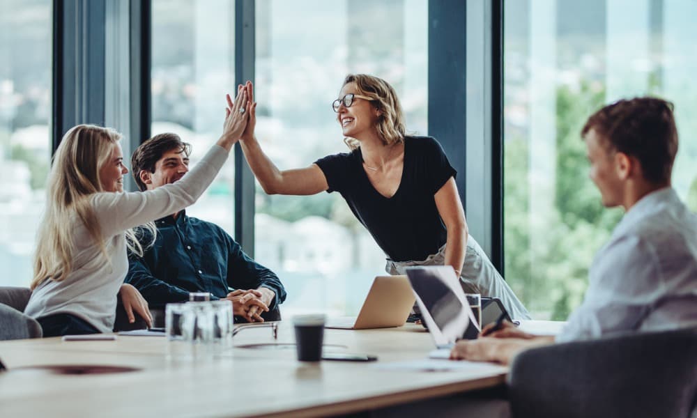 Two women giving each other a high-five at a conference table while their co-workers look on, smiling.