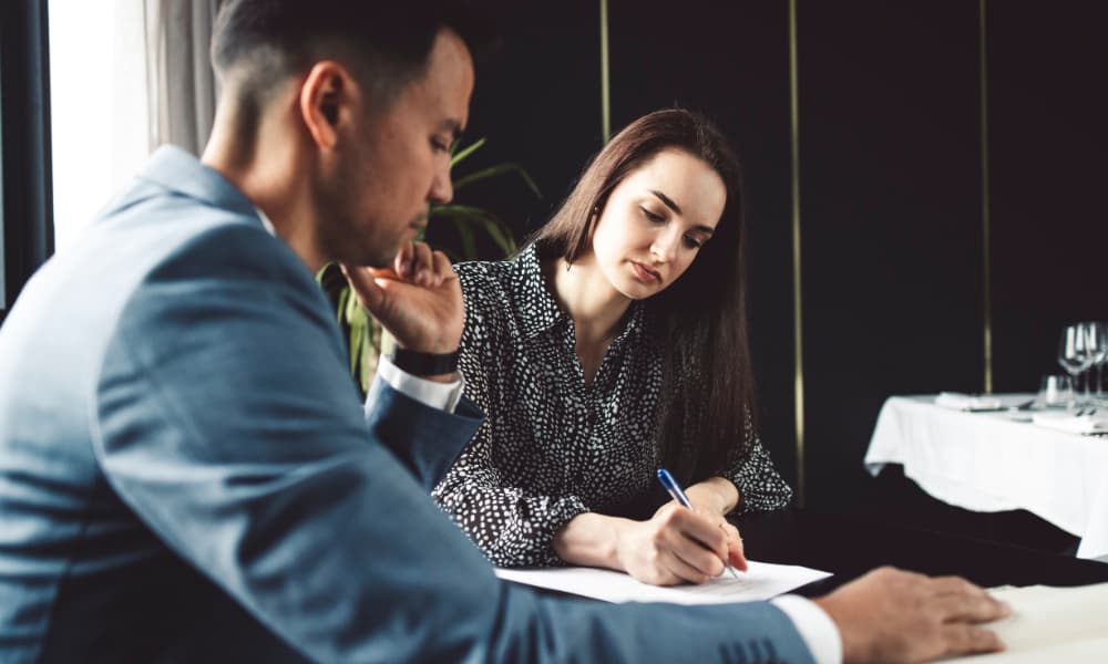 A businessman and businesswoman sitting at table while the woman signs a service level agreement (SLA)